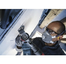 Handwerker bei der Arbeit mit einem Multifunktionswerkzeug an einer Decke, ausgestattet mit Schutzbrille und Staubschutzmaske