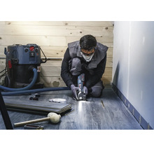 Ein Mann kniet und schneidet Laminat mit einem Multifunktionswerkzeug. Ein Staubsauger, Laminatdielen und Werkzeuge sind ebenfalls sichtbar.