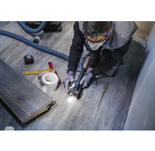 Ein Handwerker schneidet Laminat mit einer Multifunktionswerkzeugmaschine, um einen Ausschnitt für ein Rohr zu schaffen. Werkzeuge wie ein Maßband und ein Bleistift liegen bereit.