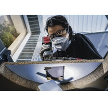 Frau mit Schutzbrille und Staubmaske arbeitet mit einem Multischleifer an einem Werkstück