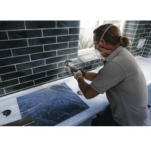 Fliesenleger bei der Arbeit mit einem Multifunktionswerkzeug an einer Wand mit schwarzen Fliesen.