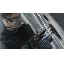 Handwerker bohrt mit einem Bohrhammer ein Loch in eine Betonwand. Er trägt dabei Schutzbrille, Atemschutzmaske und Ohrstöpsel.