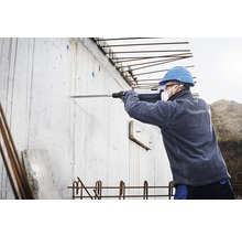 Ein Bauarbeiter bohrt mit einer Bohrmaschine in eine Betonwand. Er trägt einen Schutzhelm, eine Schutzmaske und Ohrstöpsel.