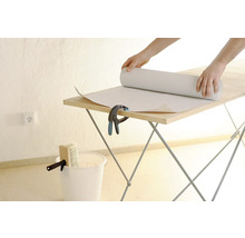 Person rollt Tapete auf einem Tapeziertisch aus. Eimer und Quast stehen bereit.