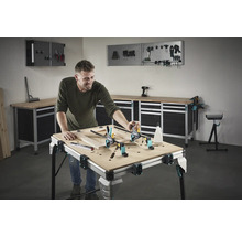 Ein Mann arbeitet an einem Holzrahmen auf einer Werkbank in einer Werkstatt. Werkzeuge und Werkstattausrüstung sind im Hintergrund sichtbar.