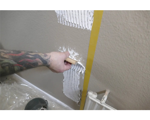 Produktbild 2 (groß) Eine Person trägt Fliesenkleber mit einer Spachtel auf eine Wand auf.