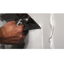 Handwerker trägt mit einer Zahnspachtel Fliesenkleber auf eine Wand auf.