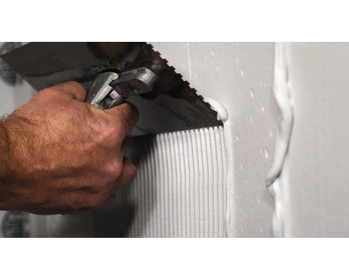 Handwerker trägt mit einer Zahnspachtel Fliesenkleber auf eine Wand auf.