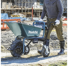 Eine Person schiebt eine mit Erde gefüllte Makita Akku-Schubkarre auf einer hellen Baustelle. Blaues Gerät mit drei Rädern und Makita Logo.