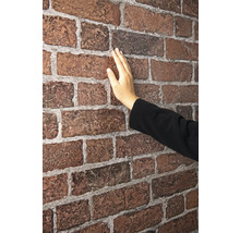 Eine Hand berührt die strukturierte Oberfläche einer Wand aus rötlich-braunen Verblendern mit hellgrauen Fugen.