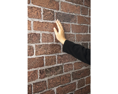 Eine Hand berührt die strukturierte Oberfläche einer Wand aus rötlich-braunen Verblendern mit hellgrauen Fugen.