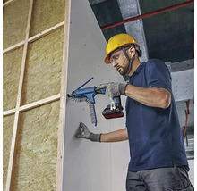 Handwerker mit Schutzhelm und Akku-Trockenbauschrauber mit Magazinvorsatz befestigt Gipskartonplatte an Holzständerwand mit Dämmung.