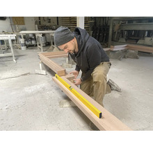 Ein Handwerker kniet in einer Werkstatt und überprüft die Ausrichtung eines großen Holzrahmens mit einer gelben Wasserwaage.