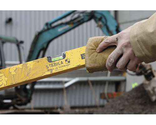 Eine Hand reinigt eine Wasserwaage mit Stabila Logo mit einem Schwamm auf einer Baustelle vor einem Bagger.