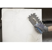 Hand mit Arbeitshandschuh trägt weißen Kleber mit einer Zahnkelle auf eine Wand auf.