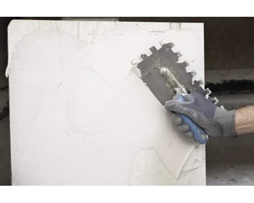 Hand mit Arbeitshandschuh trägt weißen Kleber mit einer Zahnkelle auf eine Wand auf.