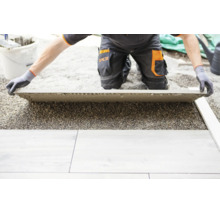 Eine Person in Arbeitskleidung verlegt eine große Terrassenplatte auf einem Splittbett.