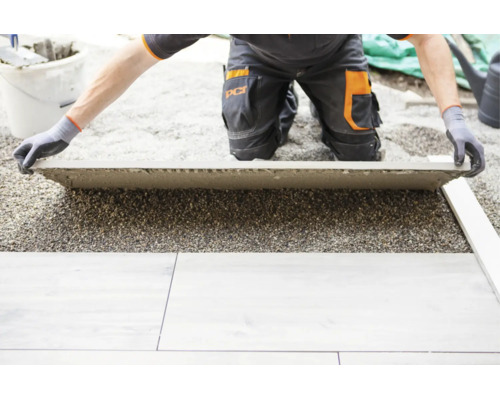 Eine Person in Arbeitskleidung verlegt eine große Terrassenplatte auf einem Splittbett.
