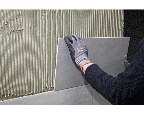 Handwerker mit Arbeitshandschuhen bringt Fliese auf mit Fliesenkleber gestrichener Wand an