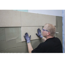 Handwerker mit Schutzbrille und Arbeitshandschuhen bringt Fliesen auf einer mit Fliesenkleber bestrichenen Wand an.