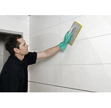 Handwerker beim Verfugen von Wandfliesen mit einem Fugengummi und Schutzhandschuhen in einem gefliesten Raum.