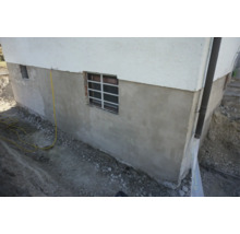 Sanierter Kellersockel eines Wohnhauses mit neuem Außenputz und Kellerschachtfenster bei laufenden Erdarbeiten.