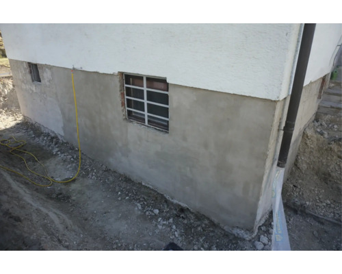 Sanierter Kellersockel eines Wohnhauses mit neuem Außenputz und Kellerschachtfenster bei laufenden Erdarbeiten.