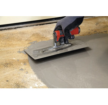 Hand in Arbeitshandschuh glättet Ausgleichsmasse auf einem Boden mit einer Glättkelle aus Metall.