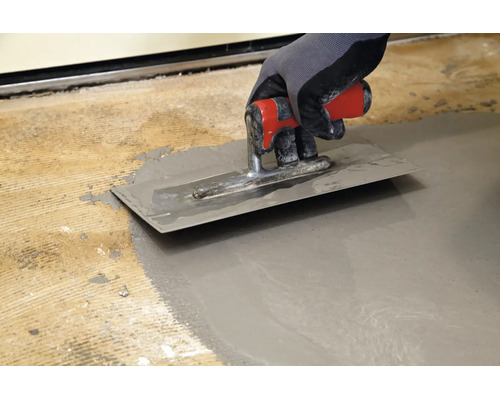 Hand in Arbeitshandschuh glättet Ausgleichsmasse auf einem Boden mit einer Glättkelle aus Metall.