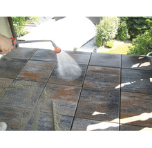 Eine Person reinigt quadratische Terrassenplatten aus Stein mit einer Sprühlanze und Wasserstrahl auf einem Balkon.