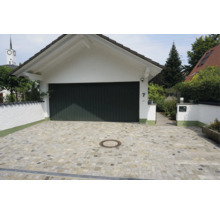 Eine gepflasterte Garagenauffahrt aus Naturstein mit einer weißen Garage und einem dunklen Garagentor vor einem Wohnhaus.