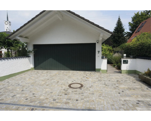 Eine gepflasterte Garagenauffahrt aus Naturstein mit einer weißen Garage und einem dunklen Garagentor vor einem Wohnhaus.