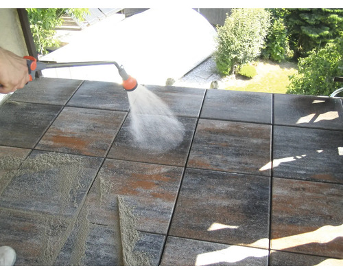 Eine Person reinigt quadratische Terrassenplatten aus Stein mit einer Sprühlanze und Wasserstrahl auf einem Balkon.