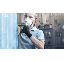 Mann schleift mit Schleifgerät eine blaue Oberfläche, geschützt mit Brille und Atemschutzmaske.