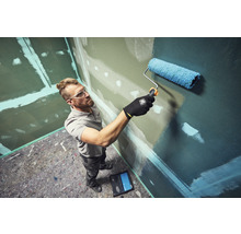 Ein Handwerker streicht eine Wand mit einer Farbrolle in einem Raum mit vorbereiteten Gipskartonplatten.