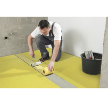 Ein Handwerker bringt mit einer Farbwalze Entkopplungsplatten auf einem Boden an. Ein Eimer mit Werkzeug steht daneben.