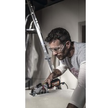 Ein Mann mit Schutzbrille schneidet mit einer Kreissäge ein Werkstück. Im Hintergrund steht eine Leiter.