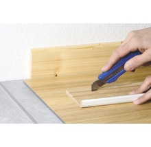 Ein Teppichmesser schneidet eine weiße Fußleiste auf einer Holzoberfläche.