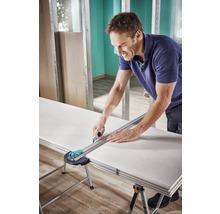 Ein Mann schneidet eine Gipskartonplatte mit einem Cuttermesser und einem Lineal auf einem Tisch.