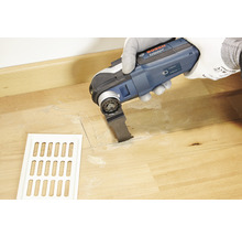 Eine Person verwendet ein Multifunktionswerkzeug von Bosch Professional, um einen Schlitz in einem Holzboden neben einem weißen Lüftungsgitter zu schneiden.