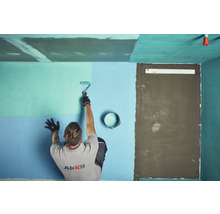 Mann streicht eine Wand mit einer Farbwalze in einem Raum. Auf dem Boden steht ein Farbeimer.