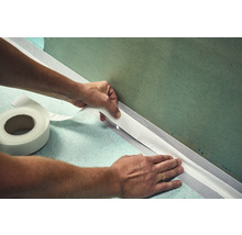 Eine Person bringt ein Dichtband an einer Wand an. Auf dem Boden liegt eine Rolle mit Dichtband.