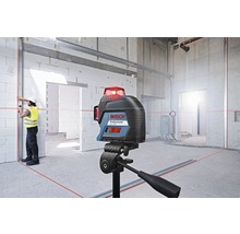 Bosch Logo. Baustelle mit Kreuzlinienlaser auf Stativ, der rote Laserlinien an Wänden und Boden projiziert. Arbeiter mit Schutzhelm und Warnweste richtet eine Metallschiene aus.