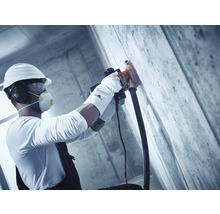 Ein Bauarbeiter mit Schutzhelm, Gehörschutz, Schutzbrille und Atemschutzmaske verwendet eine Mauernutfräse an einer Betonwand.