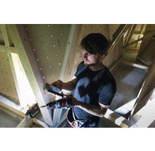 Mann fotografiert mit einem Mobiltelefon eine Bohrmaschine vor einem Holzdachstuhl