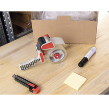 Szene mit einer geöffneten Kiste, Luftpolsterfolie, einem Klebebandabroller, einem Cutter, einem Markierstift und Notizzetteln auf einem Tisch.