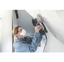 Eine Frau schleift eine Decke mit einem Exzenterschleifer und trägt dabei eine Schutzmaske sowie eine Schutzbrille.