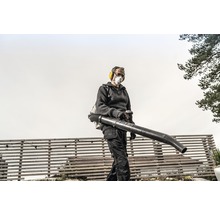 Eine Person bedient einen rückentragbaren Laubbläser im Garten, geschützt mit Gehörschutz und Staubmaske.