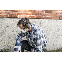 Ein Mann kniet und trägt persönliche Schutzausrüstung, darunter einen Gehörschutz, eine Schutzbrille, eine Atemschutzmaske und Arbeitshandschuhe.