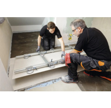 Zwei Handwerker verlegen mit einem Plattenheber Fliesen in einem Raum.